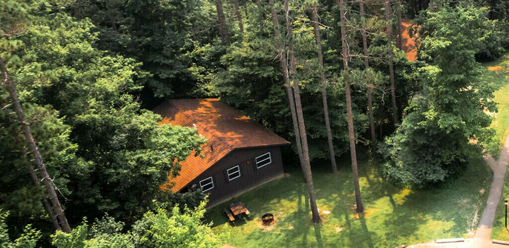 An aerial view of a grouping of timber cabins surrounded by woods at Hocking Hills State Park. Paths weave between the brown buildings and around fire pits and picnic tables. 