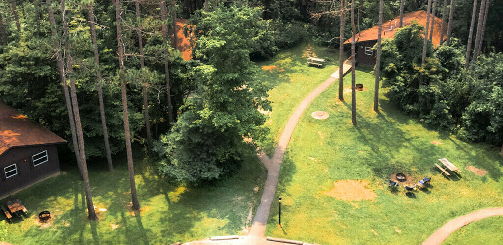An aerial view of a grouping of timber cabins surrounded by woods at Hocking Hills State Park. Paths weave between the brown buildings and around fire pits and picnic tables. 