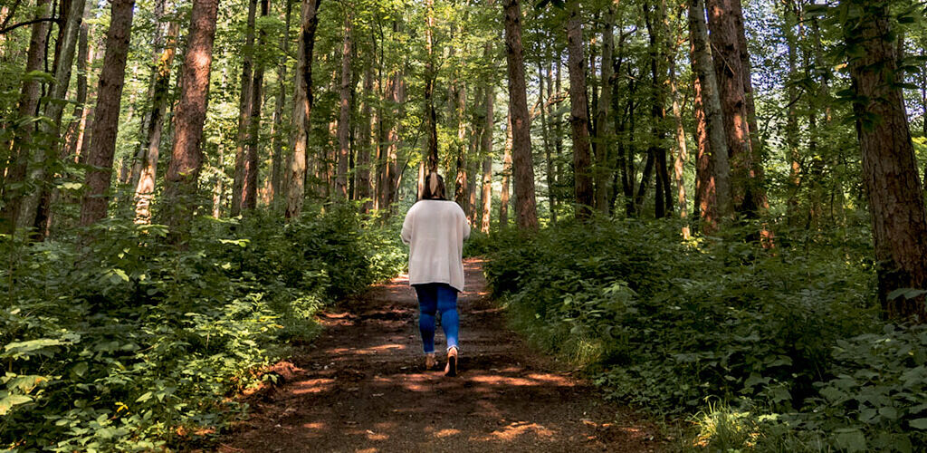 A woman walks along a dirt path through a lush green forrest. She wears blue jeans and a cream cardigan. 