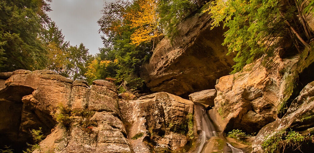 A cropping of a large rock face peeks out from among a forrest. The trees are just starting to turn from green to deeper fall colors. 